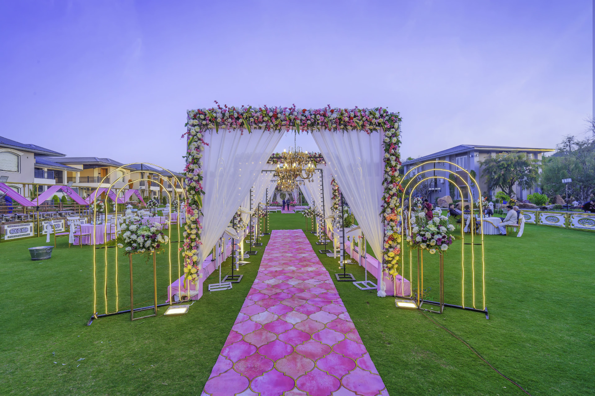 Floral mandap entrance with white drapes, chandelier and golden arches over a pink aisle