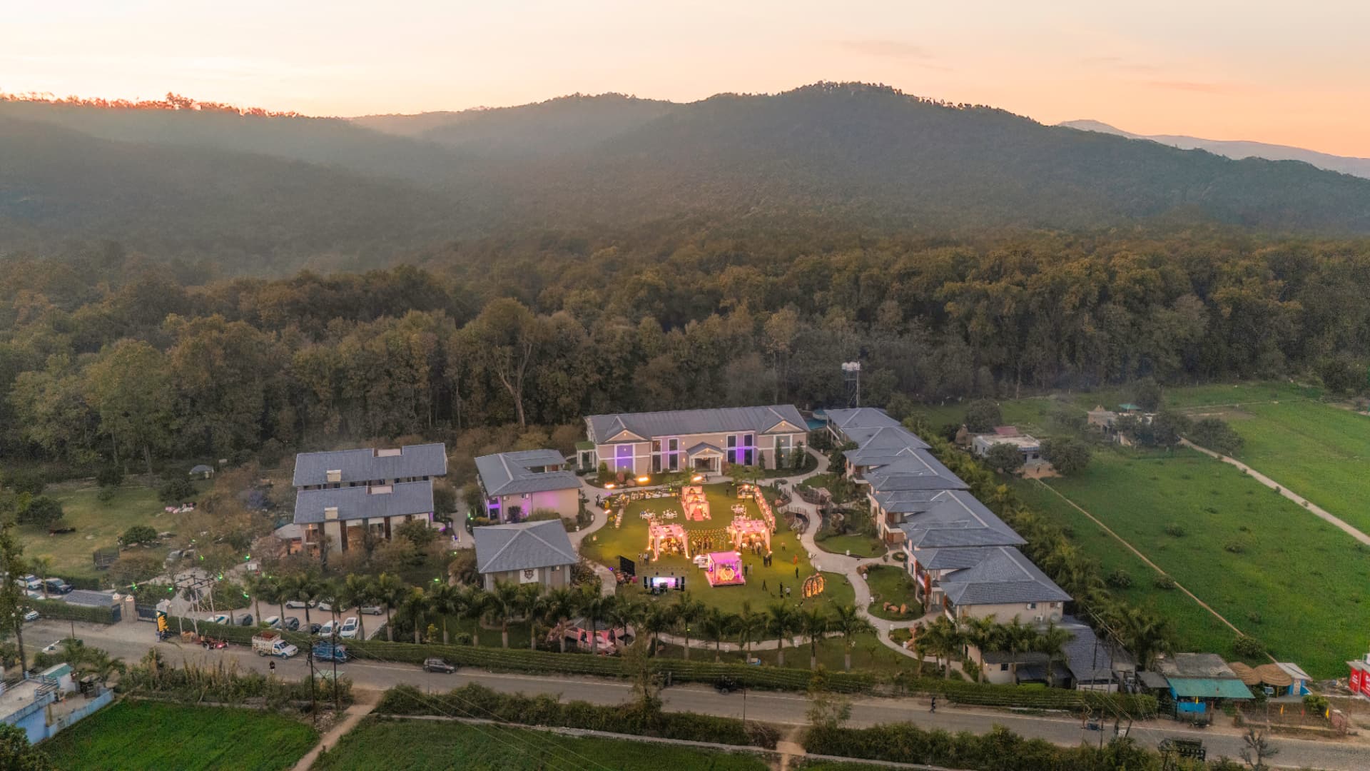 Aerial sunset view of Excel Hotels & Resorts, Kotabagh with a wedding celebration lit among villa blocks and forested hills