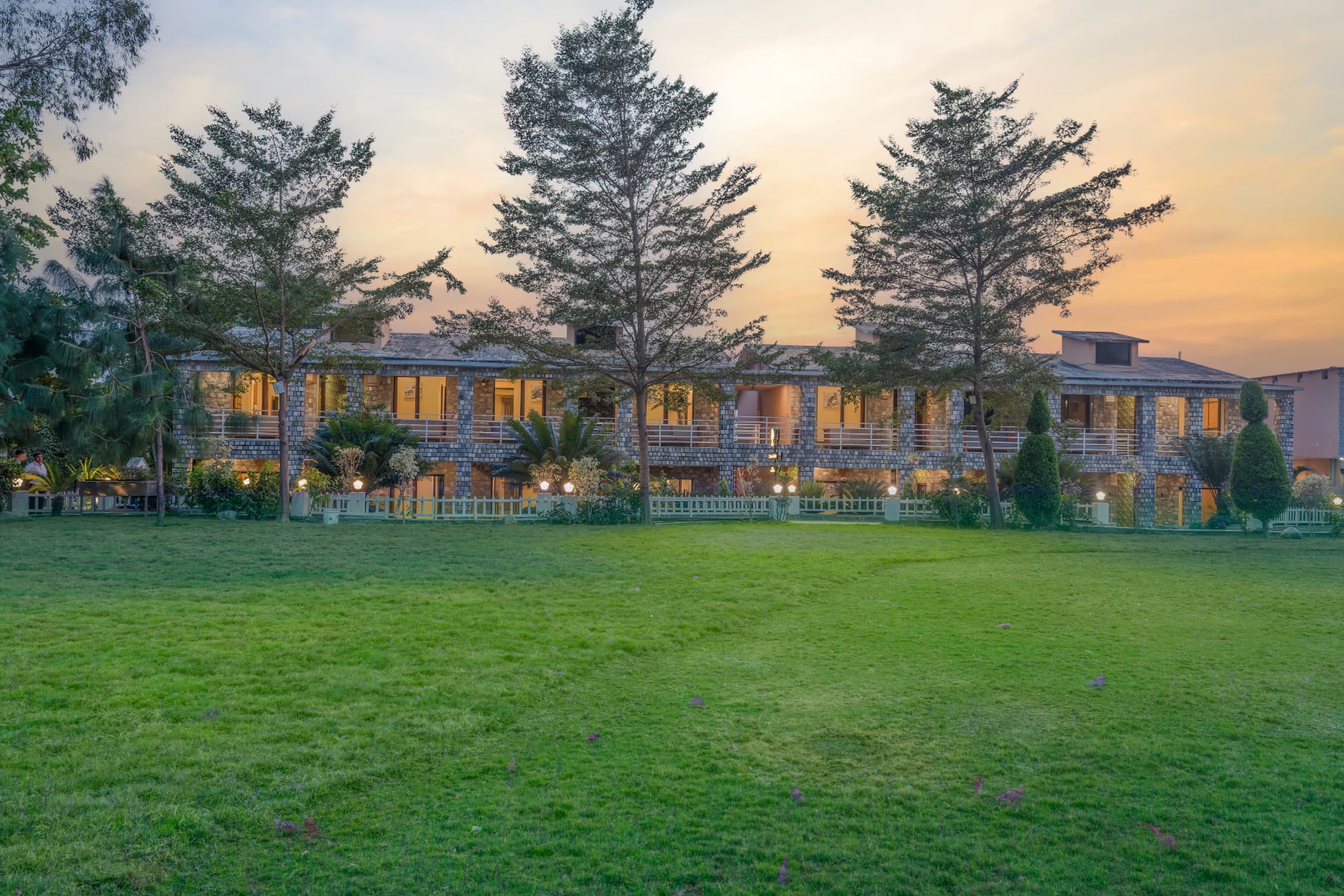 Stone-clad guest blocks glowing at sunset above the open lawn at The Banyan Retreat, Jim Corbett