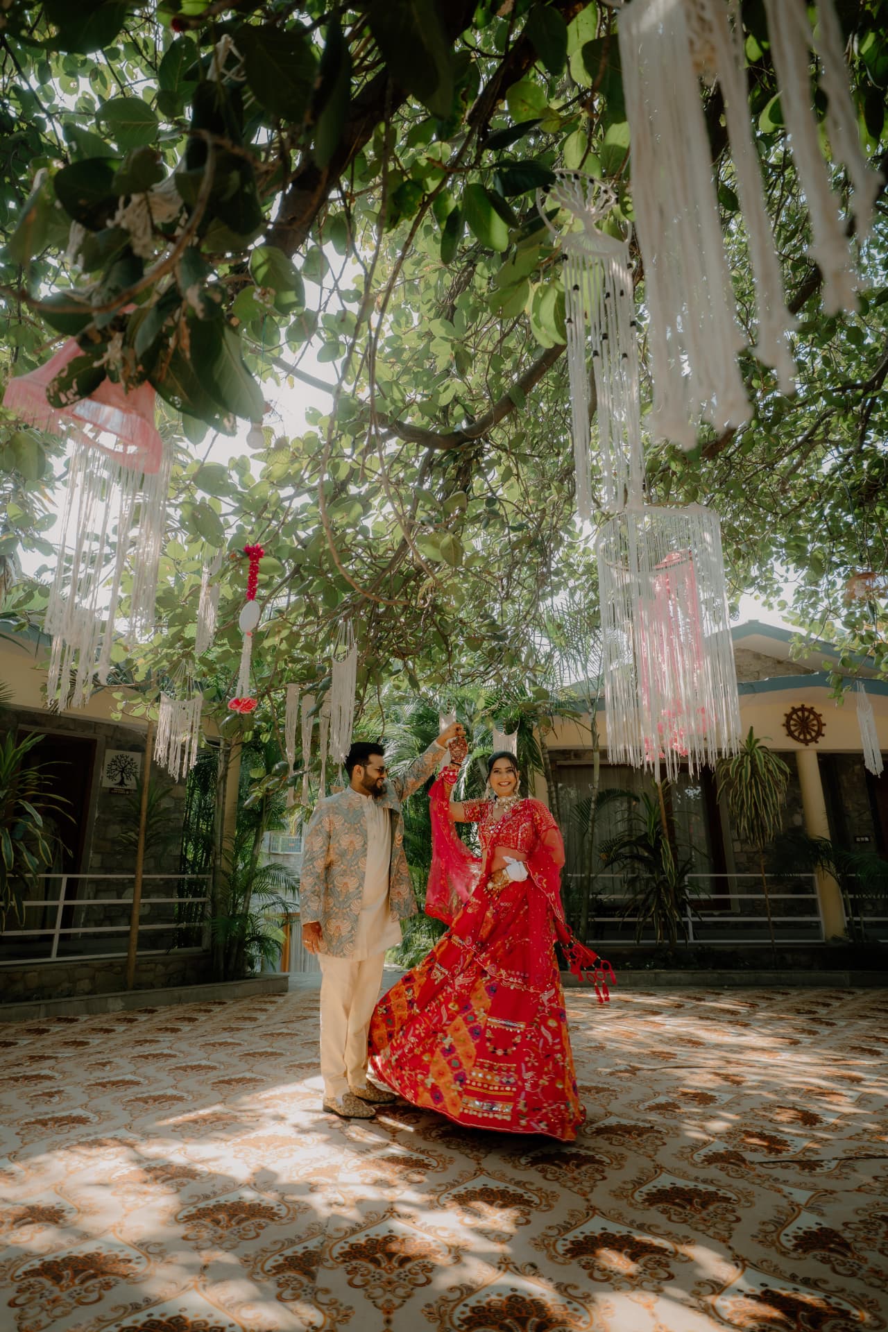 Bride in red lehenga and groom in ivory sherwani beneath hanging garden decorations at The Banyan Retreat
