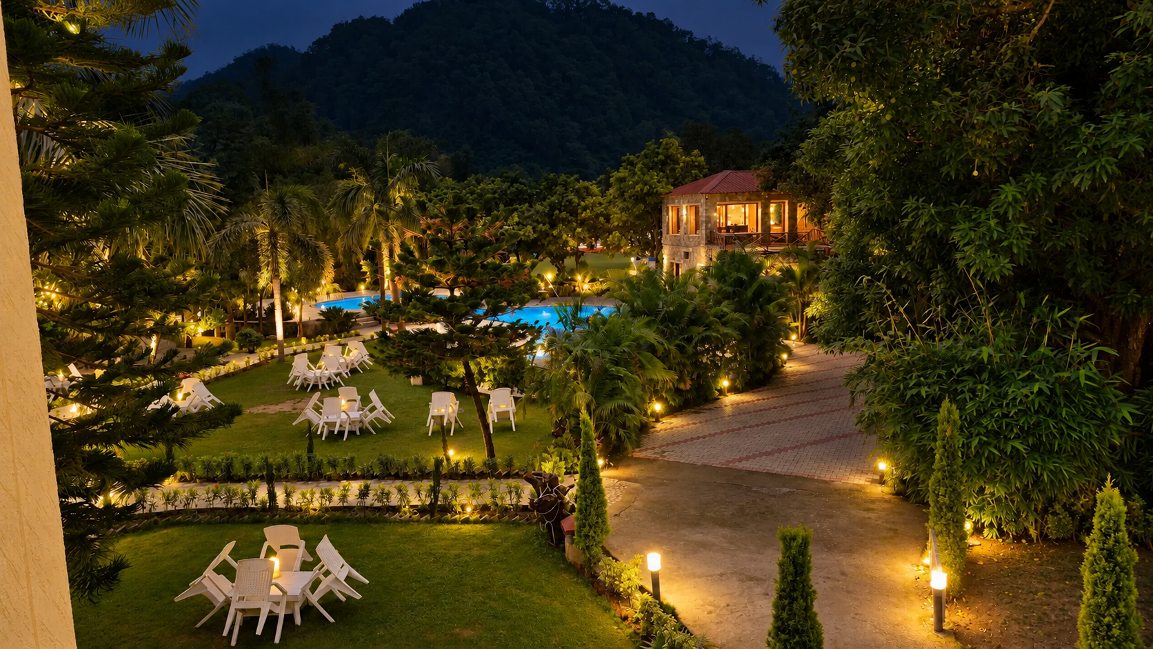 Evening view of a lit resort garden with pool, loungers and hillside greenery