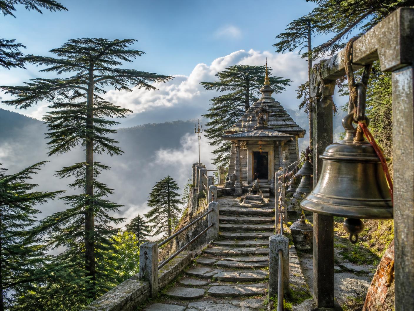 Stone steps and brass prayer bells leading to a small hilltop Shiva temple among deodar pines